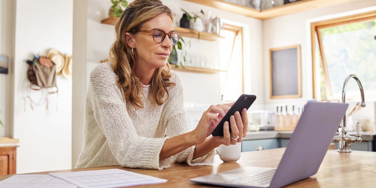 Middle aged woman sitting at her dining room table and using mobile phone and laptop while