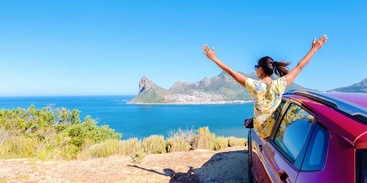 woman outside a car window with hands up, looking out over the ocean. 