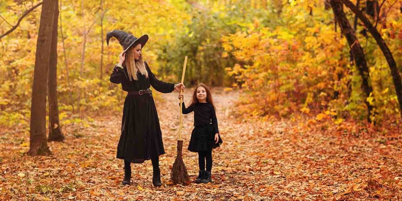 An older witch passing her broom to a young girl in the middle of a forest trail covered in leaves